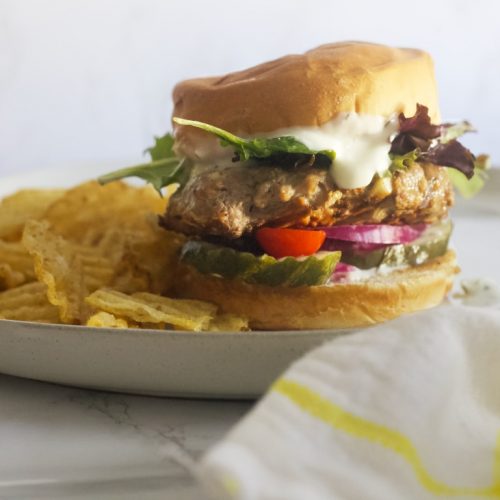 lamb burger patty with tzatziki sauce, lettuce, tomatoes, pickles, a bun and chips